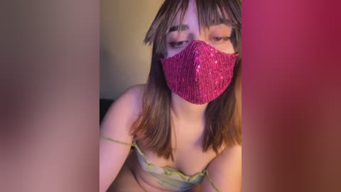 A video of a young woman with shoulder-length brown hair, wearing a glittery pink face mask and a green, strapless top, set against a blurred background of red and beige.