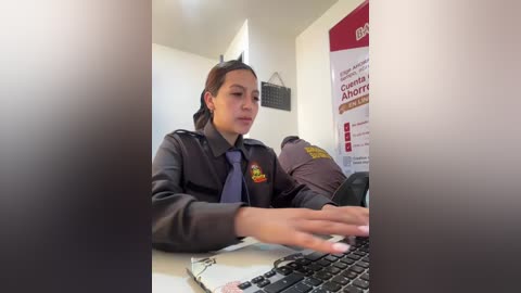 Video of a female police officer with brown hair tied back, in a dark uniform, typing on a keyboard in a small, well-lit room with a red poster on the wall.
