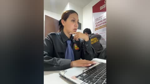 Video of a woman in a dark uniform with a blue tie, sitting at a desk, typing on a laptop, with a poster and another person in the background.