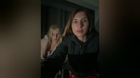 Video of two women in a dimly lit room, one in black jacket, the other in pink. They appear tense, possibly in a suspenseful scene.