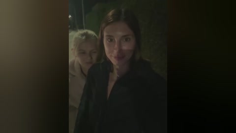Video of two women, one with platinum blonde hair and the other with dark hair, standing closely together in a dimly lit, urban outdoor setting.
