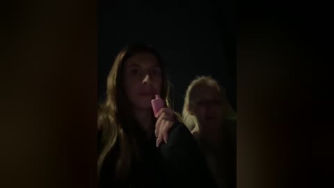 A dimly-lit video of two women, one with long brown hair and a pink lipstick, the other with blonde hair and glasses, both holding pink lollipops, creating a mysterious and intimate scene.