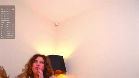 A video shows a woman with curly brown hair, wearing a brown sweater, sitting in a room with a white ceiling and a black lampshade.