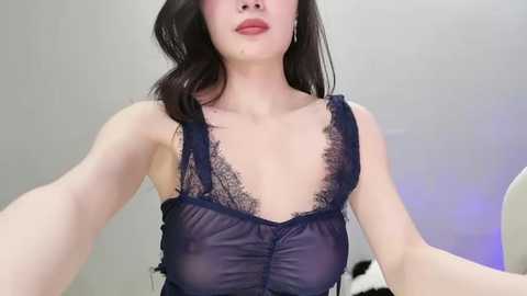 Video of a light-skinned woman with long dark hair in a sheer, dark blue lace lingerie top, revealing her breasts, in a minimalistic white room with a purple accent.