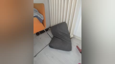 A video of a modern, minimalist bedroom with an orange chair, a gray pillow, and beige curtains, featuring a red toy on the floor.