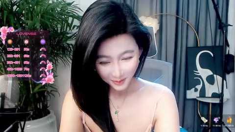 A video of an Asian woman with long black hair, wearing a pink strapless dress, smiling, in a modern room with a digital display and potted plants.