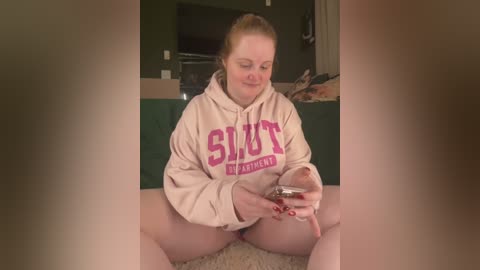 A candid video of a fair-skinned, plus-sized woman with blonde hair in a ponytail, wearing a \"SUT\" hoodie and no pants, sitting on a beige carpet, holding a wine glass, in a dimly lit living room.