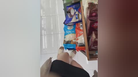 A video showing a person in black shorts bending over, grabbing a blue and white bag of rice from a white-tiled floor, surrounded by various grocery bags.
