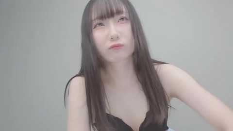 Video of a young Asian woman with long, straight black hair and light skin, wearing a black lace top, looking pensive against a plain, light gray background.