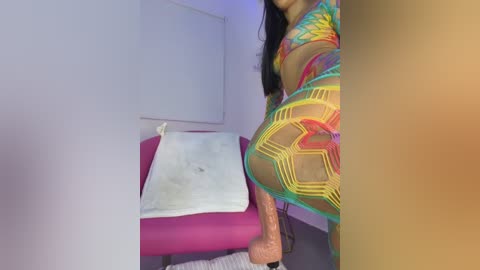 Video of a woman in a colorful, geometric-patterned bodysuit, standing beside a pink massage table with a white towel, in a minimalist, softly lit room.