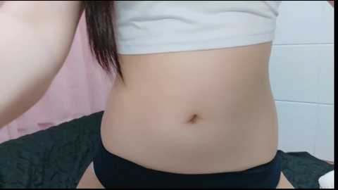 A video of a light-skinned woman's torso, showing her flat stomach, navel, and part of her upper chest. She's wearing a white crop top and black panties. The background features a pink wall and a green bedspread.