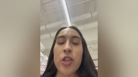 Video of a young Asian woman with long black hair, fair skin, and full lips, making a kissy face, taken from a low angle in a bright, industrial room with white ceiling and exposed pipes.
