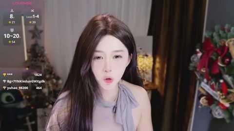 Video of an East Asian woman with long, straight black hair, wearing a light gray halter top, in a room decorated with holiday ornaments and a wooden chair.