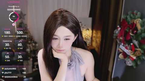 A video of an Asian woman with long, straight, dark brown hair, wearing a gray sleeveless dress, sitting indoors with a pensive expression.