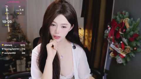 A video of a young Asian woman with long black hair, pale skin, and a slender build, wearing a sheer white blouse, looking contemplative in a dimly-lit room decorated with Christmas ornaments.