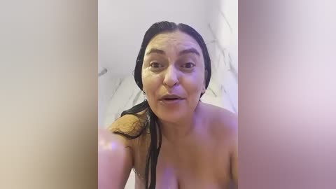 Video of a woman with wet hair, wearing a beige towel, taken from a low angle, in a bathroom with a tiled floor.
