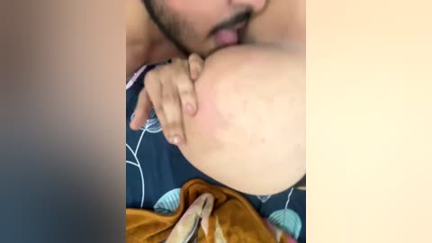 A video shows a close-up of a man with a beard sucking on a woman's large breast. She has a fair complexion and is wearing a blue patterned bra.