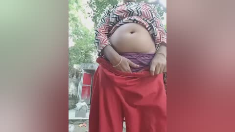 A video of a woman with a medium complexion, wearing a patterned top and red pants, partially unbuttoned, standing in front of a red building with greenery in the background.