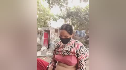 Video of a woman with dark hair, wearing a face mask and a patterned jacket, in a rural outdoor setting with makeshift structures and trees in the background.