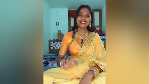 Video of a smiling Indian woman in an orange and yellow sari, adorned with gold jewelry, sitting on a bed in a modest room with white walls and wooden furniture.