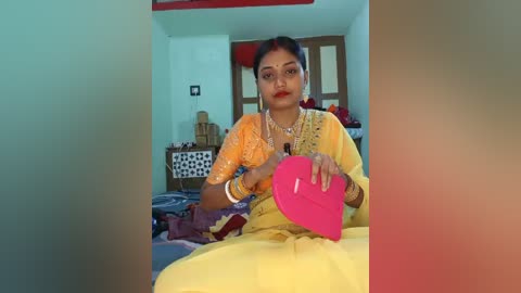 Video of a South Asian woman in a bright yellow sari, seated on a bed, holding a pink object. The background features a simple room with a wooden door and storage boxes.