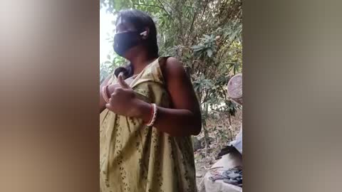 A video of a dark-skinned woman with long black hair, wearing a green floral dress, black mask, and a pink bracelet, giving a thumbs-up, in a lush, green, outdoor setting.