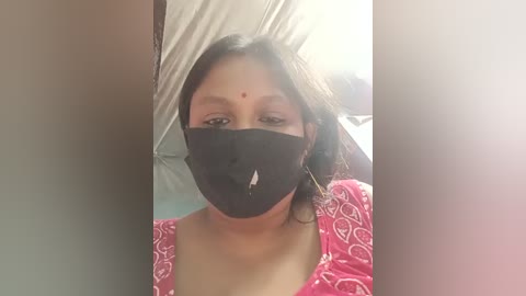 Video of a woman with medium brown skin, wearing a black mask, pink floral-patterned blouse, and a red bindi. She stands in a tent-like structure with white fabric.