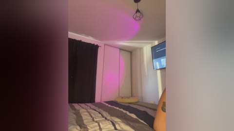 A video of a dimly-lit, sparsely furnished bedroom with a black curtain, a bed with a patterned duvet, a TV on the wall, and a small yellow pillow on the floor.