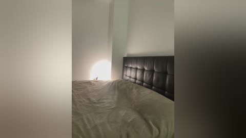 A video of a minimalist bedroom featuring a black, tufted headboard against a plain white wall. The bed is made with white, wrinkled sheets. A soft, white lamp glows dimly on the nightstand.