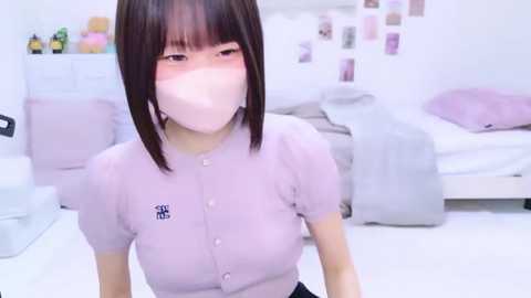 Video of an East Asian woman with straight black hair, wearing a pink cardigan and face mask, in a pastel-themed bedroom with a white bed and pink accents.