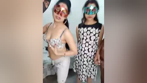Video of two Asian women with long black hair, wearing reflective, multicolored masks and matching floral-patterned dresses. One is in a bra, the other in a skirt, standing indoors.