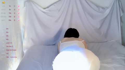Video of a woman in white lingerie, lying face down on a bed with white sheets, partially covered by white drapes.