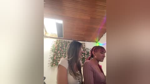 Video of two women in a dimly lit room with a wooden ceiling, one with long brown hair, wearing a white top, and the other with red hair, in a pink sweater.