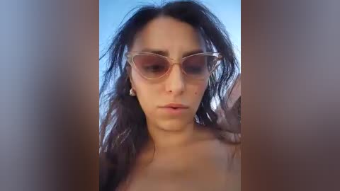 Video of a young woman with long, dark hair and fair skin, wearing oversized, clear-rimmed sunglasses, standing against a blurred, neutral background.