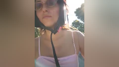 A video of a young woman with light skin, wearing a pink camisole, sunglasses, and a pink choker, standing outdoors with blurry trees and a clear sky in the background.