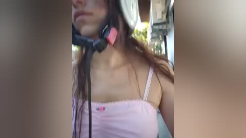 Video of a young woman with long, dark hair, wearing a pink spaghetti strap top and a black headset, taken indoors with blurred background.