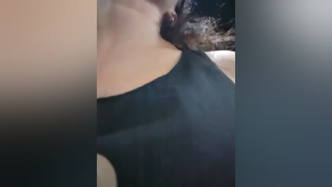 A video showing a close-up of a person's neck and shoulders, wearing a black, sleeveless top. The background is blurred, with a dark, textured surface. The person has curly hair, partially visible.