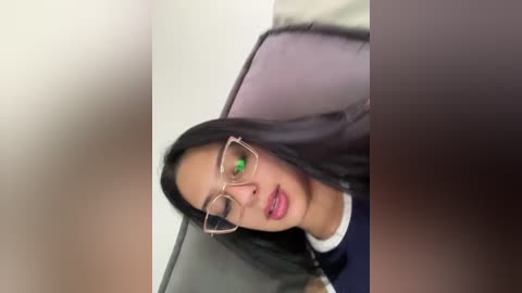 Video of a young woman with long black hair, wearing glasses and a blue top, lying on a gray pillow against a beige wall, with a blurry background.