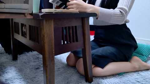 Video of a person, likely female, kneeling on a gray shaggy rug, wearing a white sweater and dark skirt, typing on a wooden desk with a laptop and books, in a well-lit room.