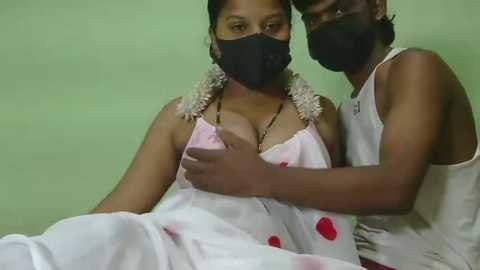 Video of an Indian couple in a dimly lit room; woman wearing a white polka-dotted dress, black mask, and flower garland, man in a white tank top, both with dark skin.