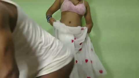 Video of a woman in a white dress with red heart prints, holding the skirt, wearing a pink bra, against a green wall.