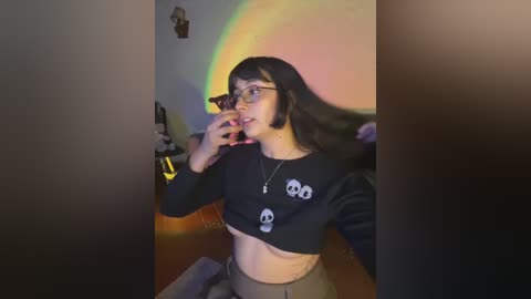 A video of a young woman with long black hair, glasses, and a black crop top with skull designs, taking a selfie in a dimly lit room with a vibrant rainbow projected on the wall.