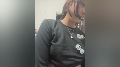 Video of a woman with a black bandana over her mouth, wearing a black sweatshirt with skull prints, standing in a dimly lit room.
