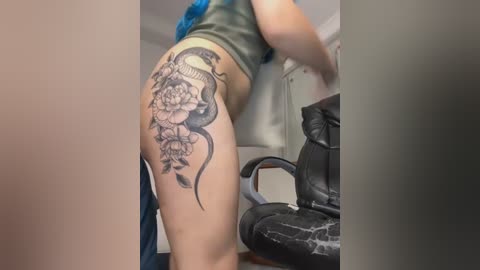 Video of a person with a large, intricate black and grey tattoo on their left thigh, featuring a snake and floral design. They are wearing a green crop top and blue hair, standing in a room with a black office chair.