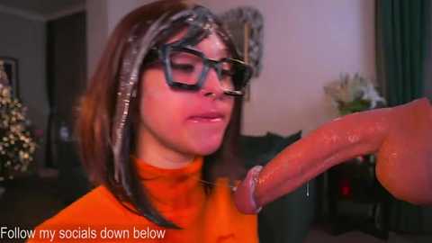 Video of a woman with shoulder-length brown hair, wearing glasses, an orange turtleneck, and a glittery headband, receiving a large erect penis, in a dimly lit room.
