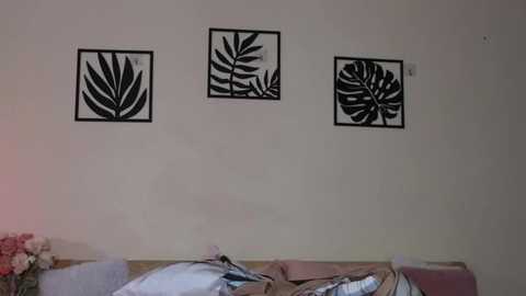 Video of a beige wall with three black-and-white leaf-shaped wall decals in a grid pattern. A light pink bed with a white comforter and a small bouquet of pink flowers on the left.