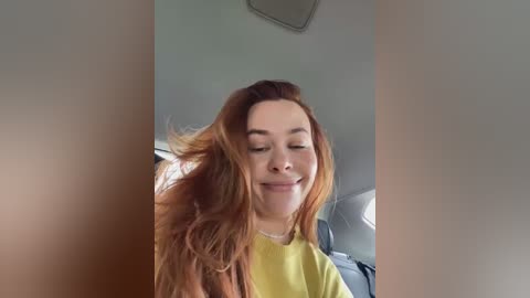 Video of a smiling young woman with long, wavy, auburn hair, wearing a yellow shirt, captured from the neck up, in a car.