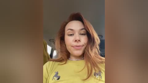A video of a fair-skinned woman with long, wavy red hair, wearing a yellow T-shirt with horse motifs, posing with a slight pout in a dimly lit room.