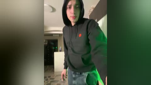 A video of a young woman with a slender physique, wearing a black hoodie and jeans, standing in a dimly lit hallway with green lighting. Her expression is serious.