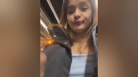 Video of a young woman with long brown hair, wearing a blue tank top and black jacket, leaning against a car door with a blurred background.